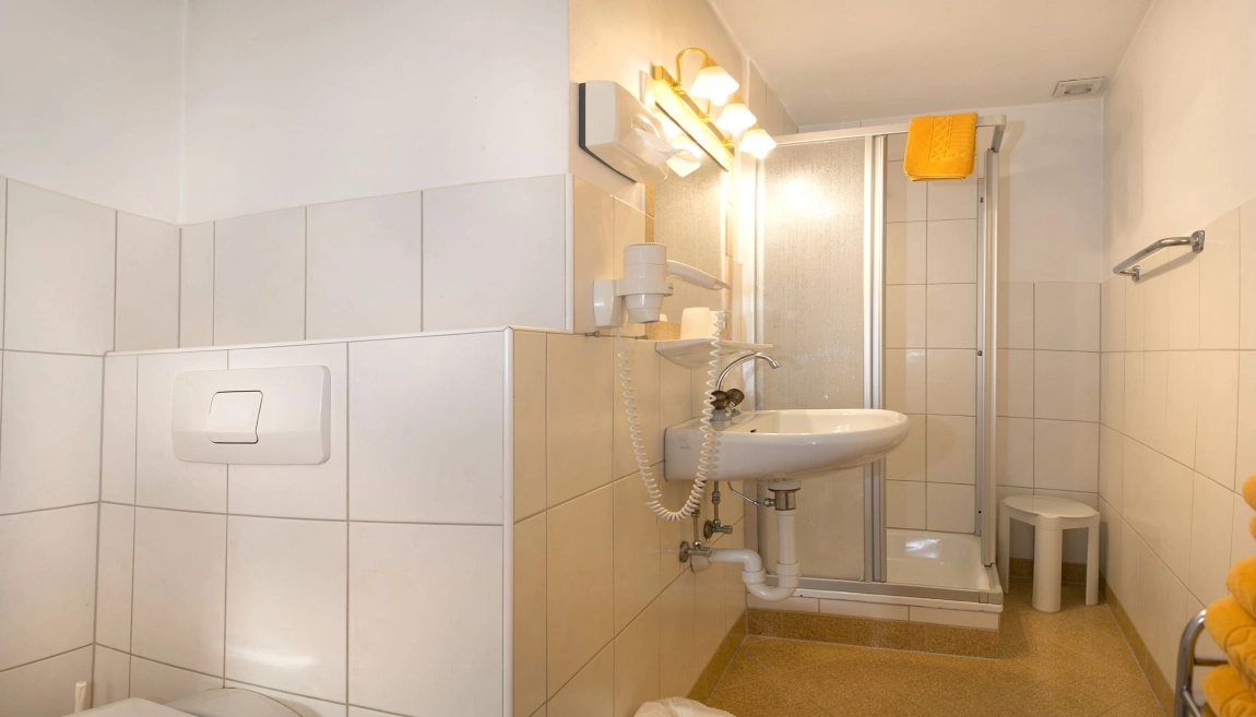 Helles Bad mit weiß gefliesten Wänden, Dusche mit Glaswand, WC, Waschbecken und gelbem Handtuch auf Haken.